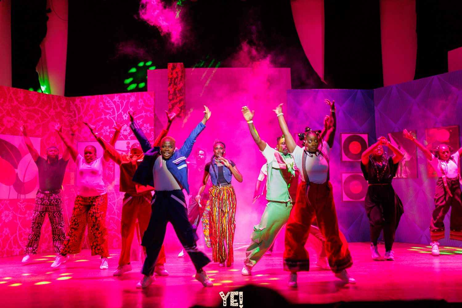 The YE! company with arms raised in pink stage light