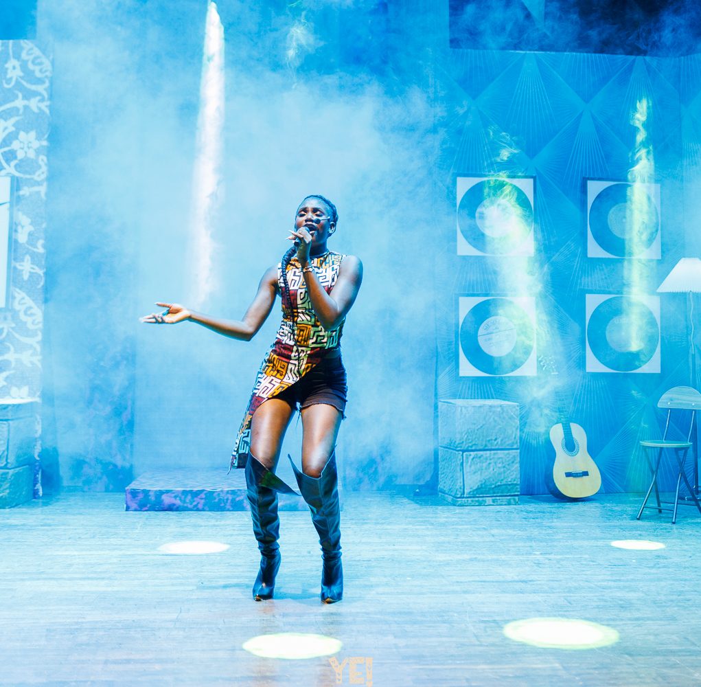 A YE! performer sings in a patterned dress and tall boots